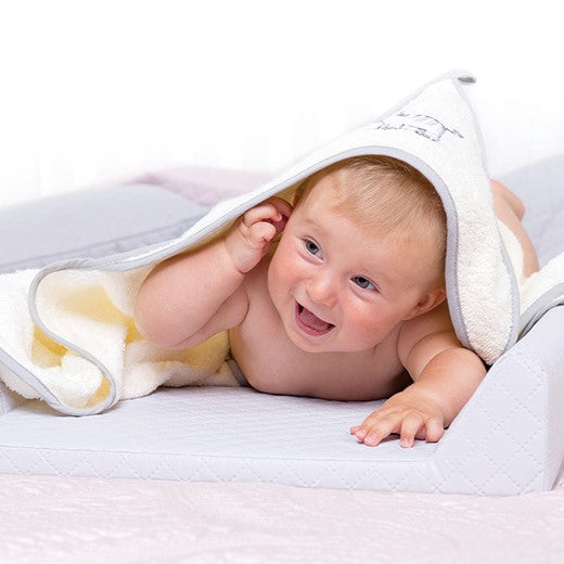 Young smiling baby laying on their stomach with a hooded bath towel over their head on top of a white padded Ceba Baby changing mat.